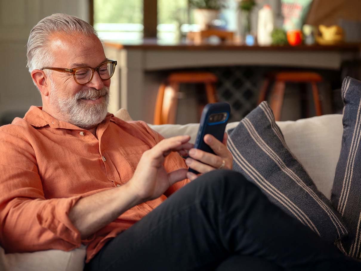 A smiling man relaxing on his couch, checking his hearing and wellness stats on his phone